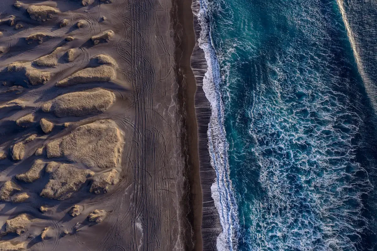 Top-down aerial view of Iceland’s black sand beach with ocean waves breaking along the shoreline and textured sand dunes shaped by wind and water.