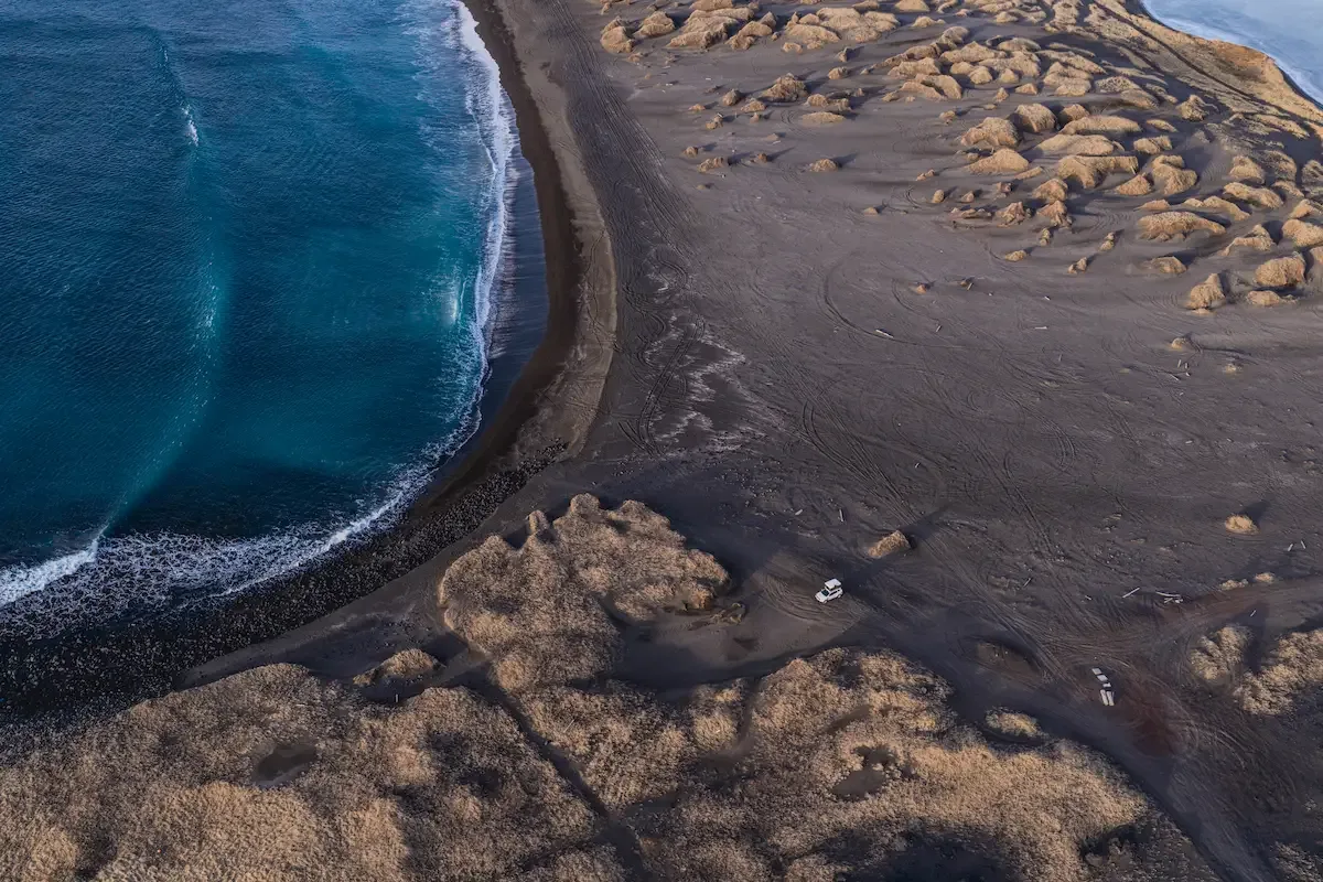 Aerial view of Iceland’s coastline with black sand beach, turquoise ocean waves, and a campervan parked near the shore during a scenic road trip.