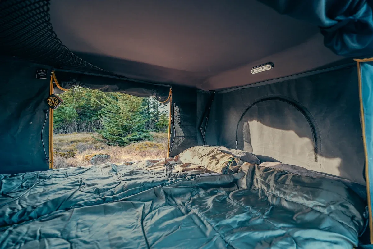 Interior of a campervan sleeping area in Iceland, showing a comfortable bed with bedding and an open rear door looking out to a natural landscape.