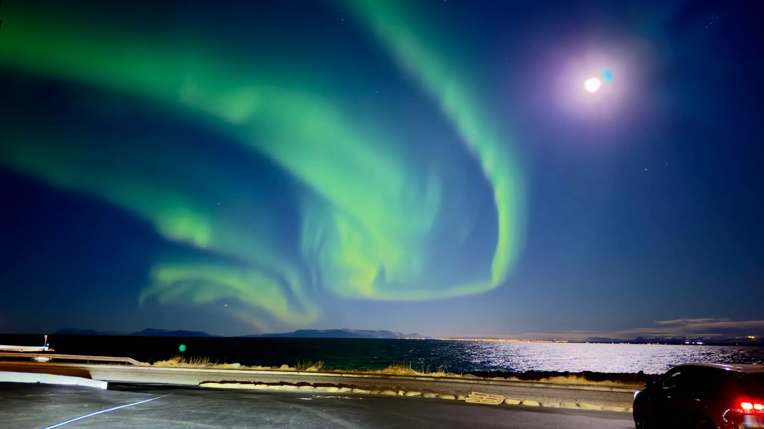 Intense green and red Northern Lights in Iceland illuminating the night sky above travelers parked along a dark roadside.
