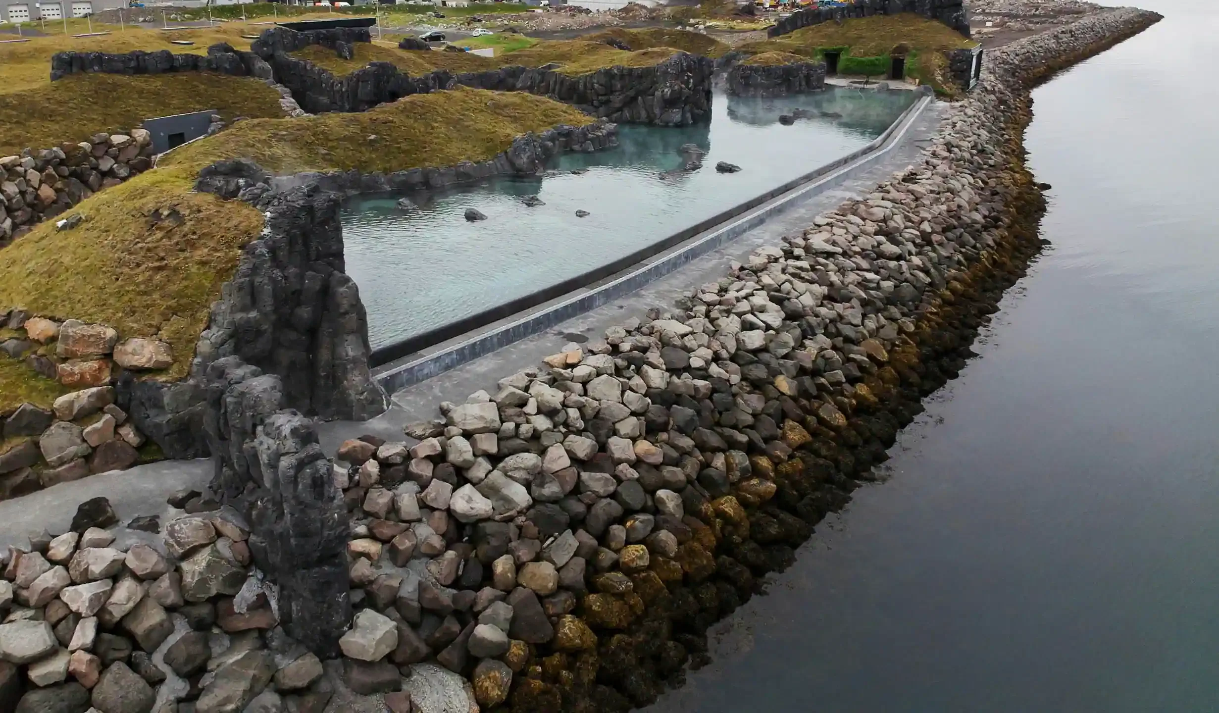 An Icelandic geothermal pool with bright turquoise mineral water enclosed by ancient basalt lava formations covered in green moss and reinforced with a rocky stone breakwater