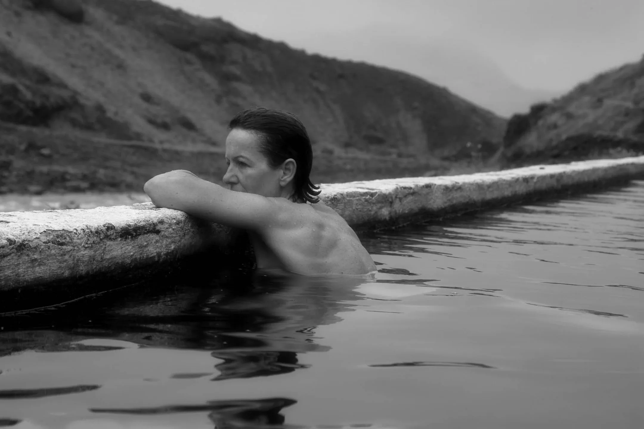 A traveler relaxing in a natural geothermal hot spring surrounded by dramatic mountain peaks and remote Icelandic wilderness in monochrome