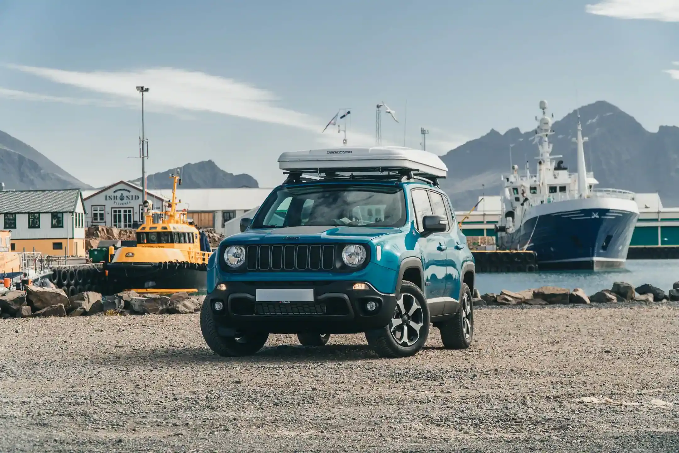 Jeep with roof tent parked near an Iceland harbor with boats and mountains in the background