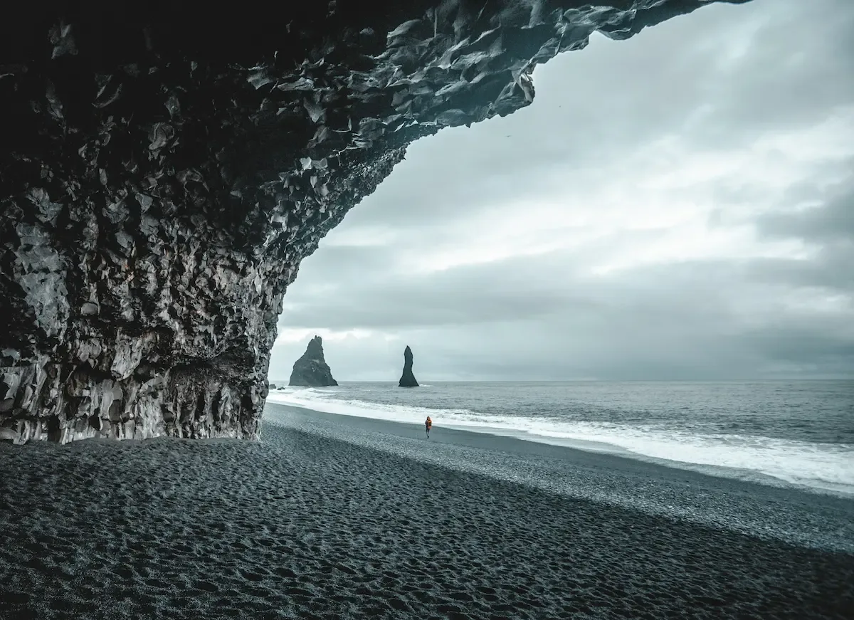 View from inside the basalt cave at Reynisfjara Black Sand Beach, looking out toward the Reynisdrangar sea stacks and the Atlantic Ocean under cloudy skies.