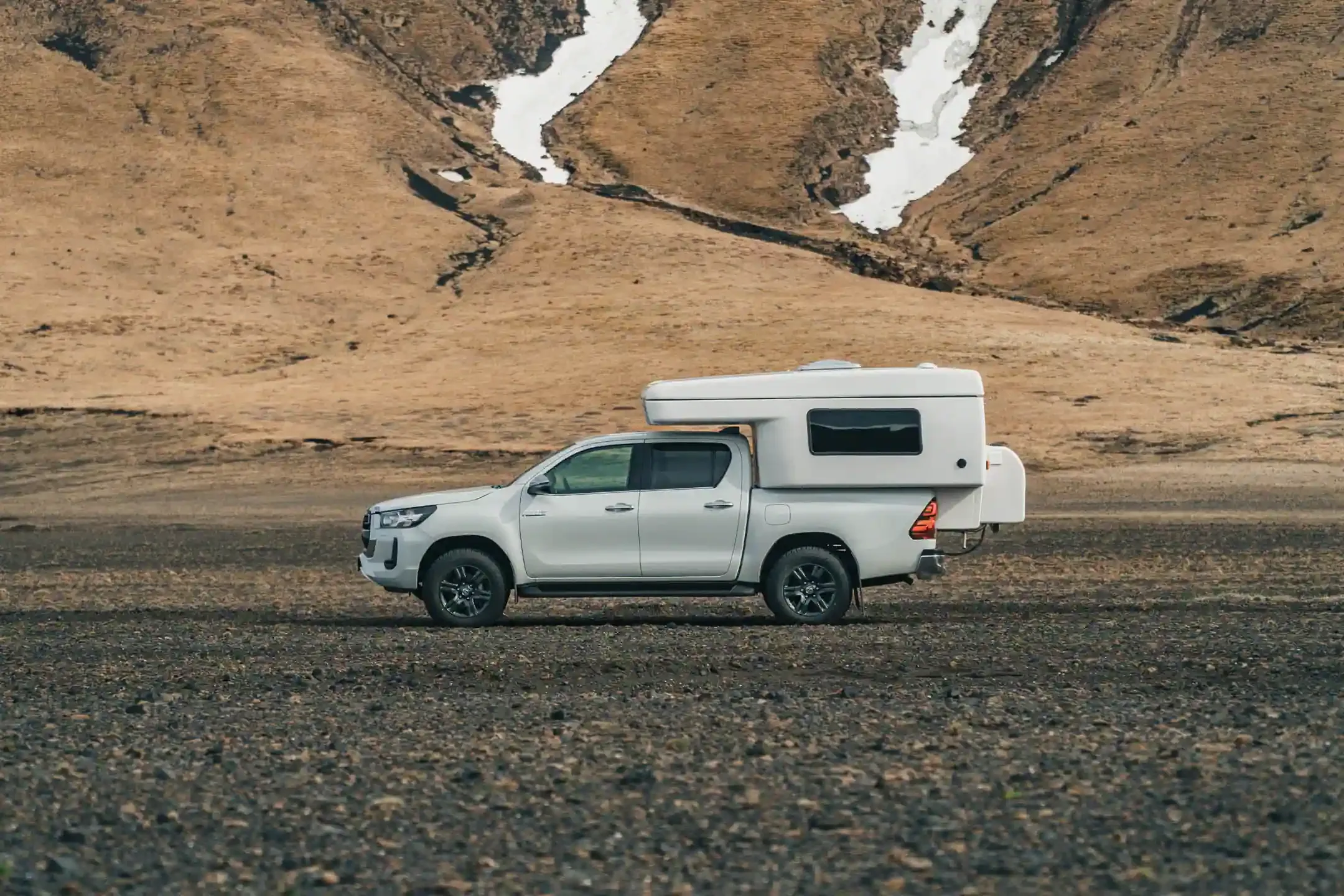 Toyota Hilux 4x4 camper driving through the Icelandic highlands with snow patches on surrounding mountains.