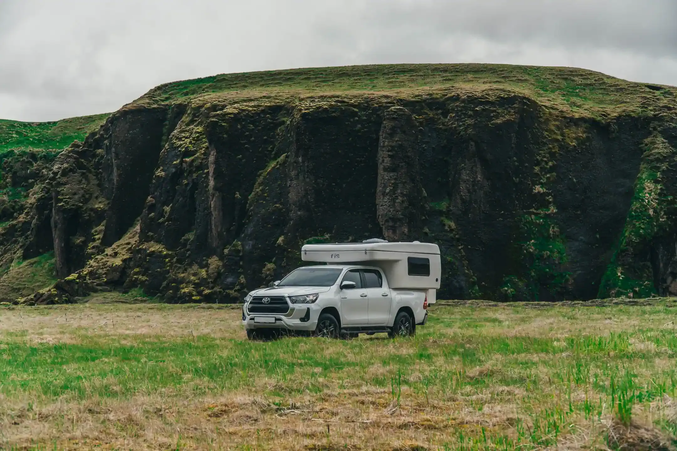 White Toyota Hilux 4x4 campervan with camper shell parked in front of green moss-covered cliffs in Iceland
