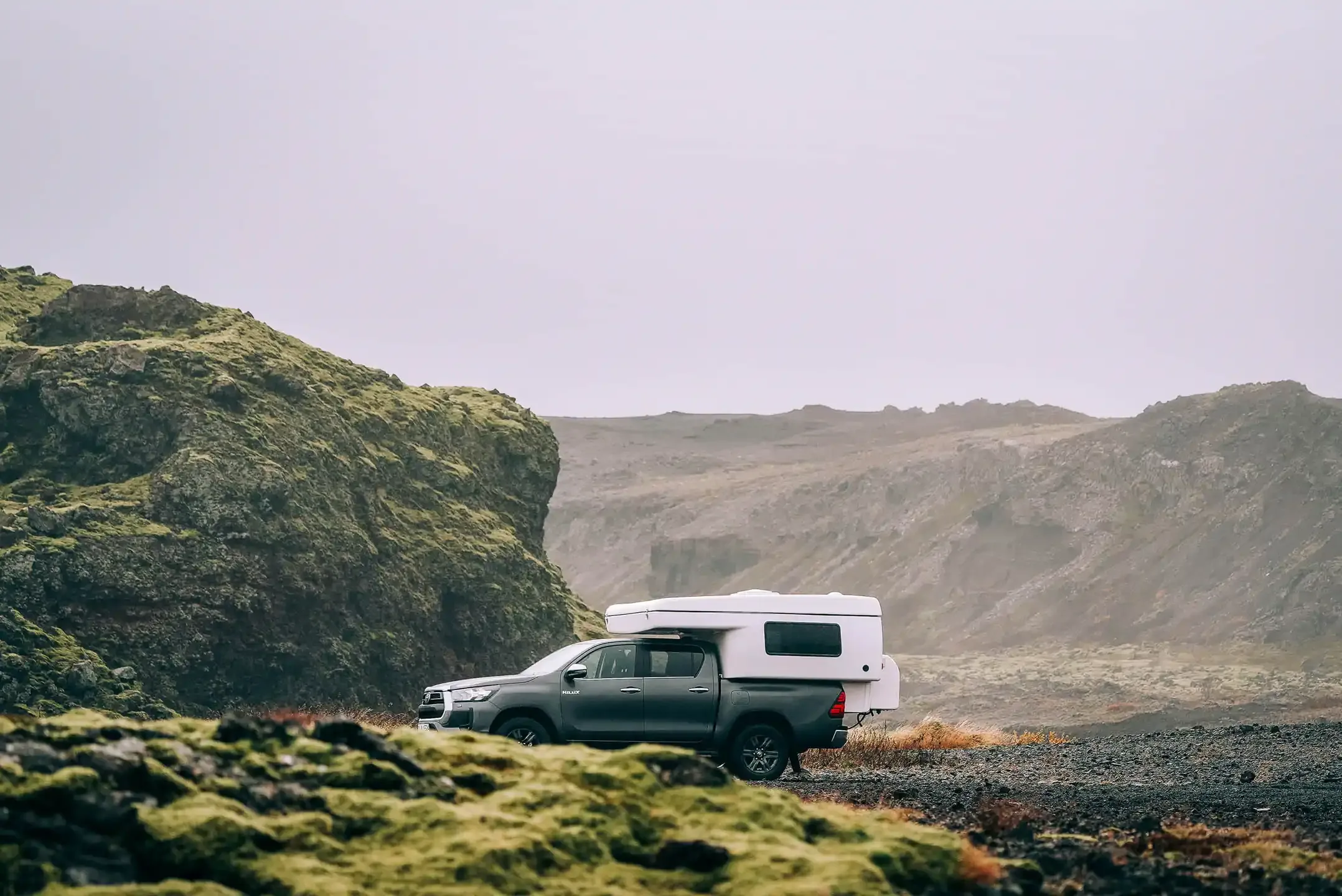 White Iceland campervan parked beside a mossy lava field and calm lake on a cloudy winter day, showcasing remote travel scenery in Iceland