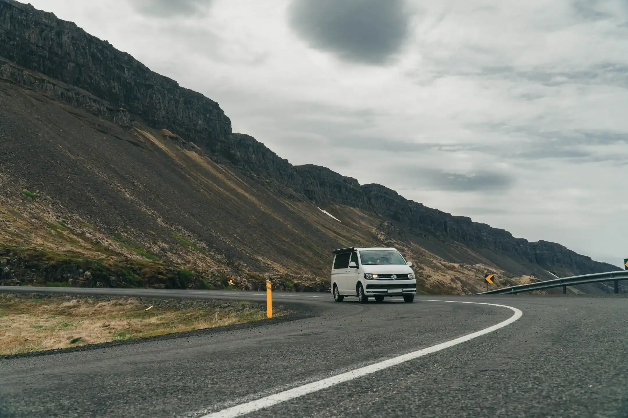 Family campervan Iceland driving along a winding coastal mountain road with dramatic cliffs and cloudy skies