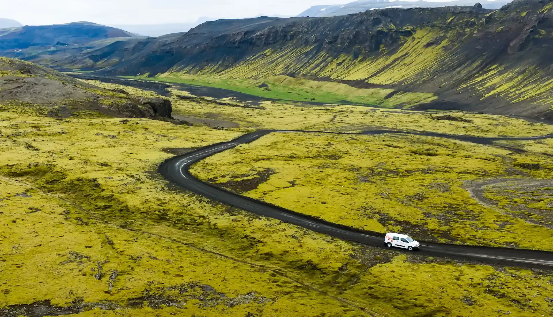 A campervan driving along a winding mountain road through moss-covered volcanic landscapes in Iceland's remote Westfjords