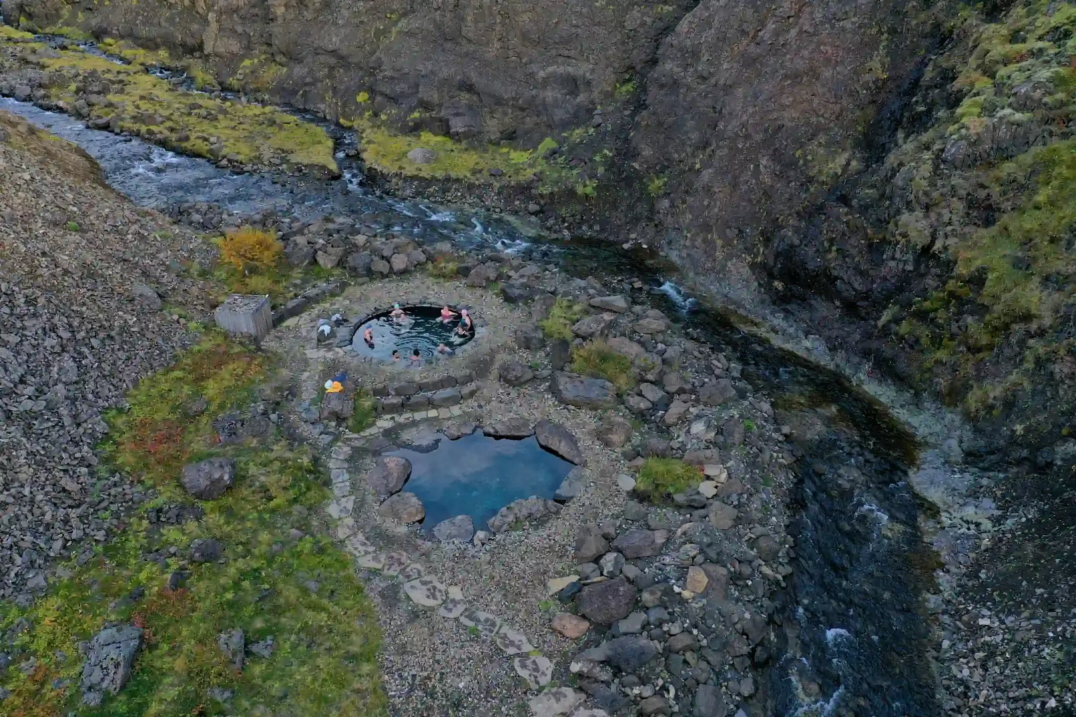 Aerial view of natural stone hot spring pools in Iceland beside a river in a rocky valley.