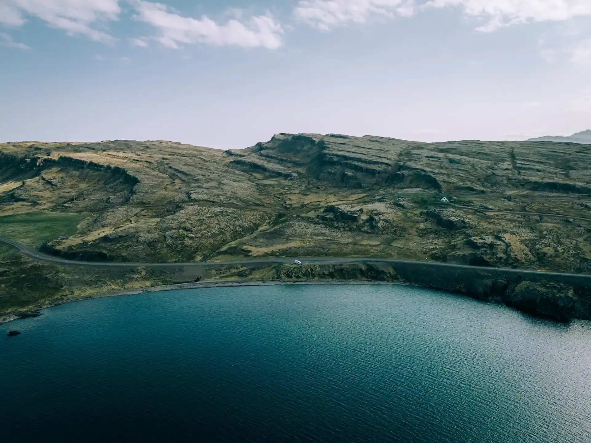 A stunning turquoise glacial lake in Iceland surrounded by green moss-covered volcanic lava landscape with rolling mountains and a winding road visible at the top