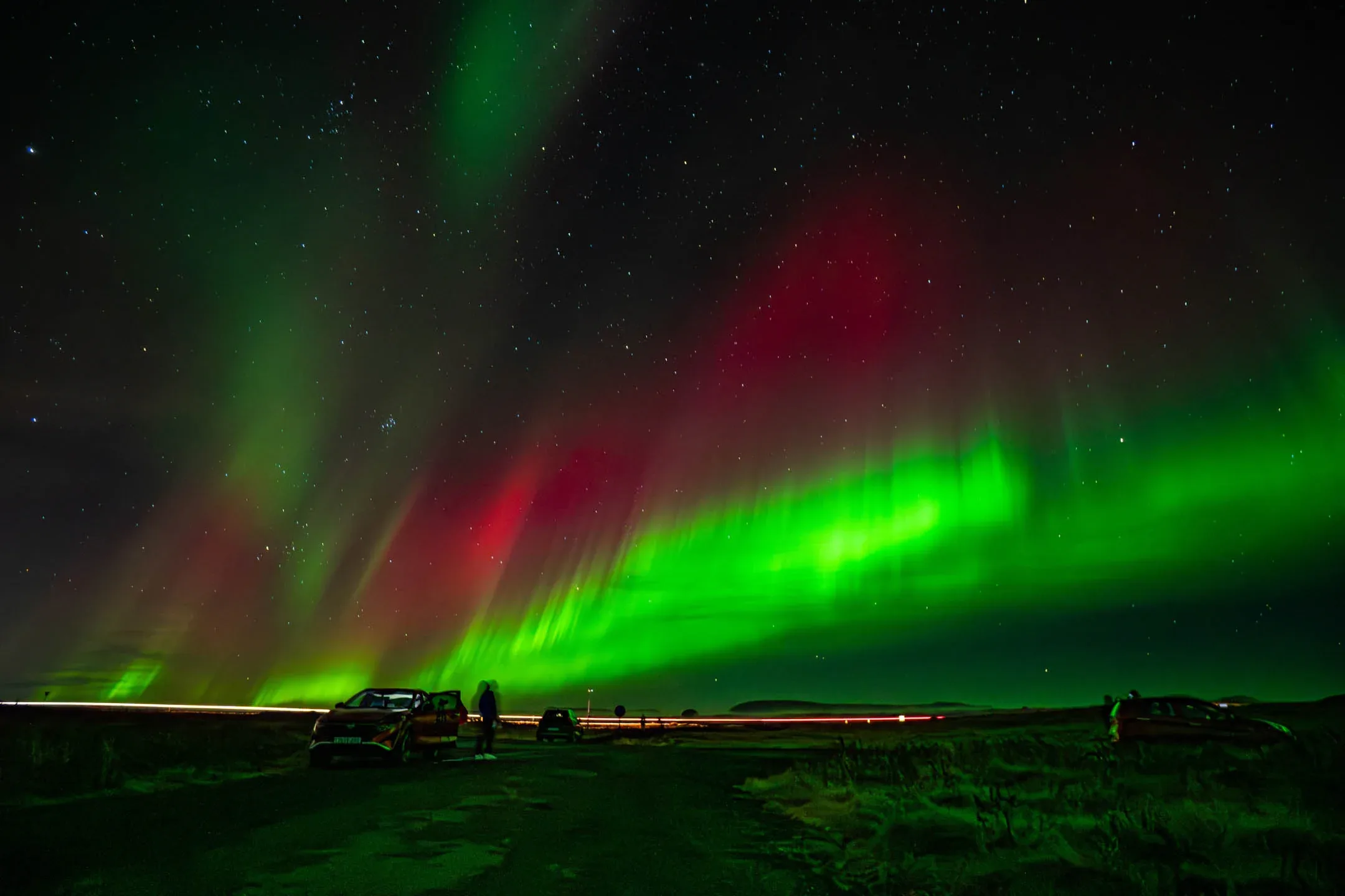 Bright green and red Northern Lights over a dark road in Iceland with parked cars and stars in the sky