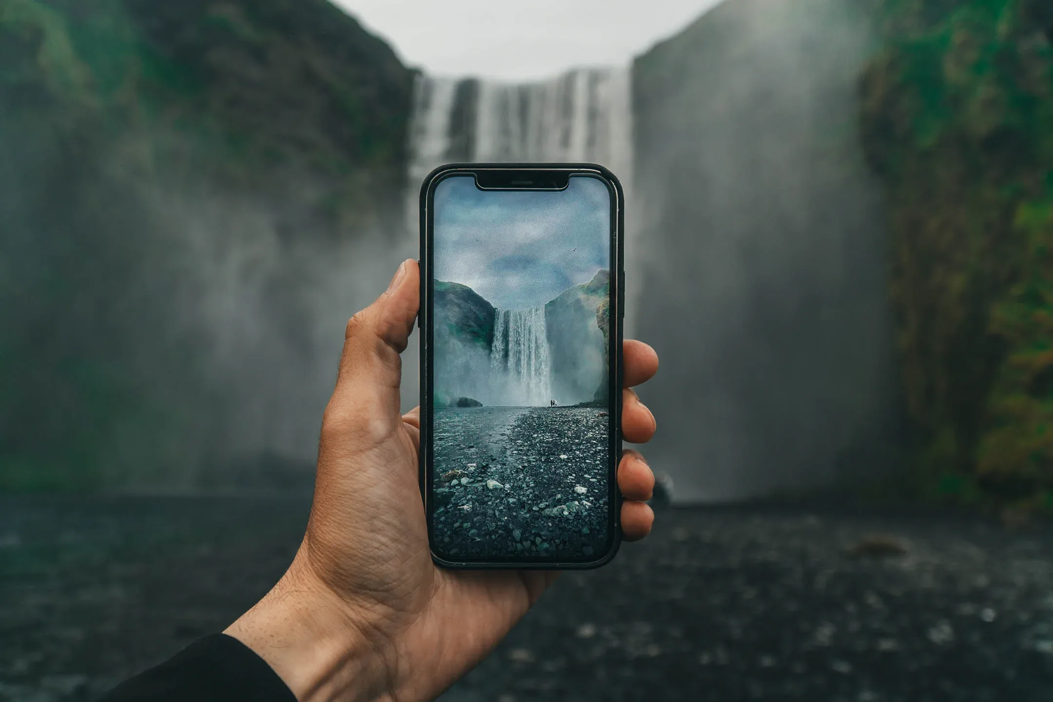 Hand holding smartphone framing Skógafoss waterfall, with misty falls and rocky riverbed in southern Iceland