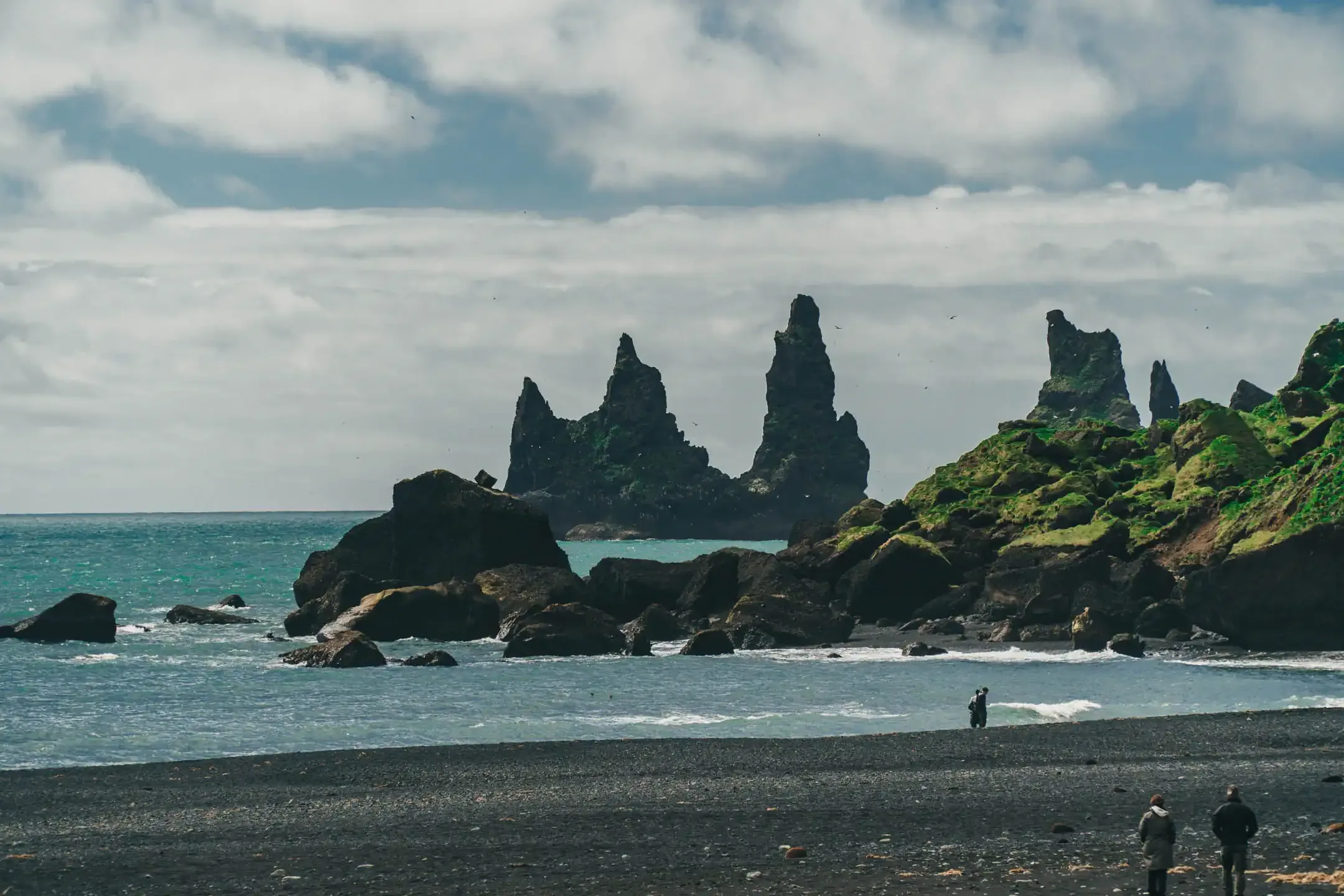 Sea stacks of Reynisdrangar rising from the ocean off Reynisfjara black sand beach in South Iceland, with moss-covered rocks and tourists on the shore