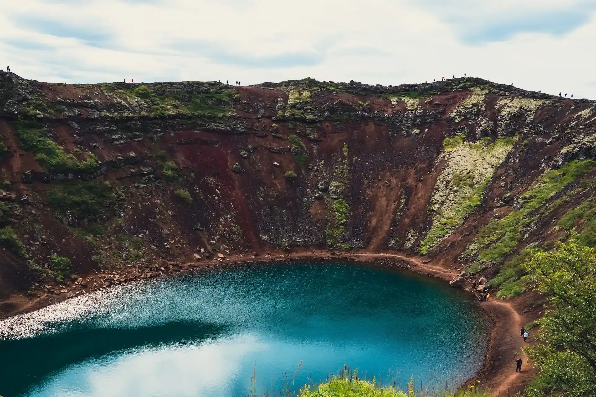 Kerið volcanic crater lake in Iceland, with vivid blue water surrounded by red volcanic rock and green moss.