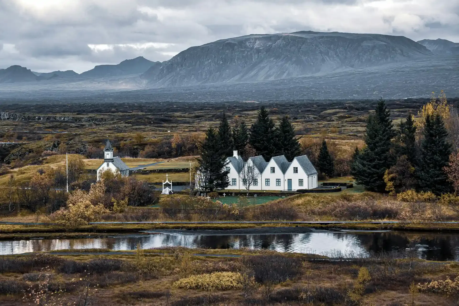 Þingvellir landscape with a white church, historic buildings, autumn trees, and mountains beside a calm lake.